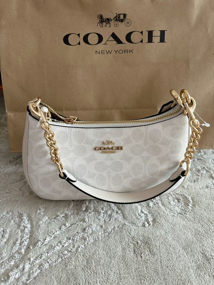 A Coach hobo shoulder bag in signature monogram canvas, featuring a gold-tone chain strap and a white leather shoulder strap, displayed in front of its brown Coach New York shopping bag.